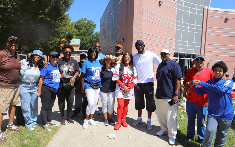 A group of people standing together outdoors, smiling in front of a brick building with large windows.
