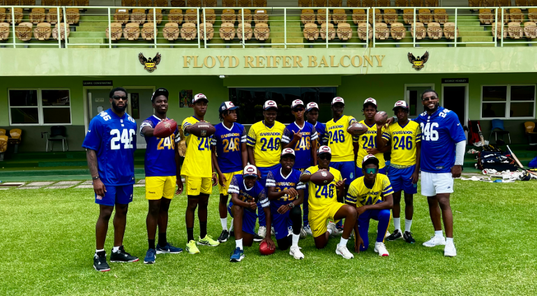 A group of young athletes in blue and yellow sports uniforms smiles on a green field. They're holding footballs, exuding a sense of teamwork and joy.