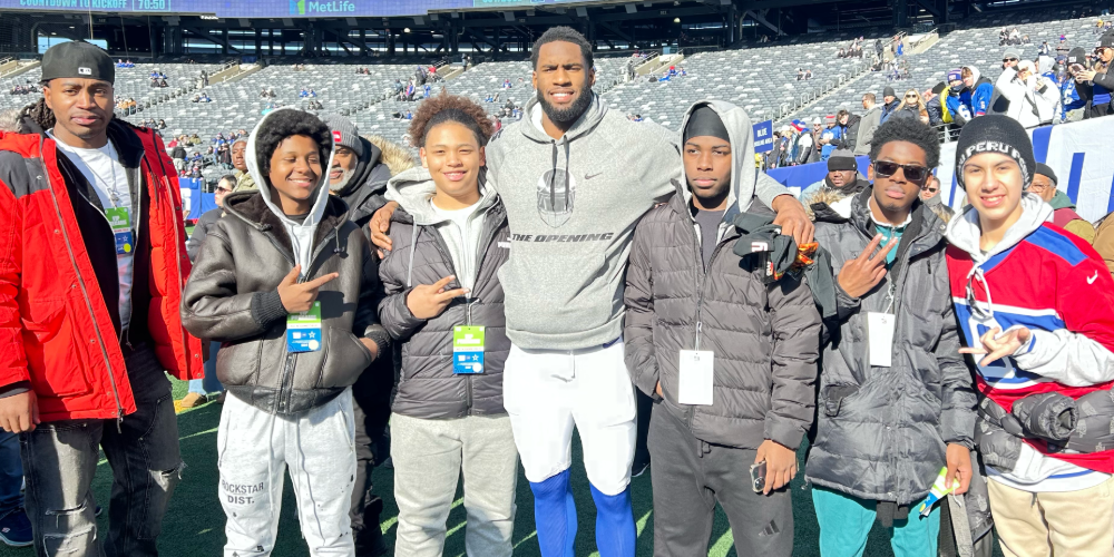 A group of seven people, dressed in casual and warm clothing, smile on a sunny day in a stadium. They appear cheerful and are posing together.