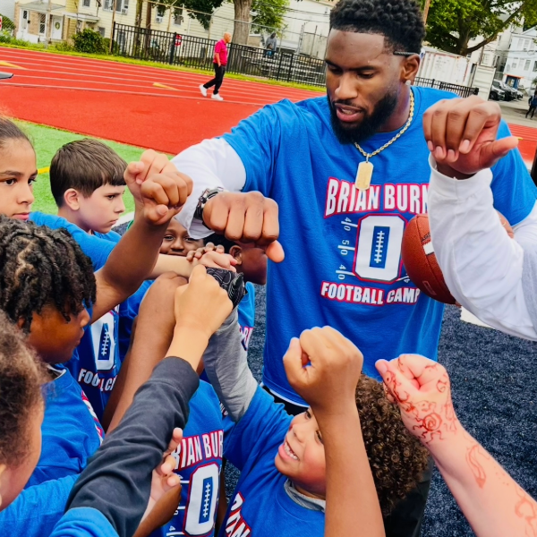 A group of children and a man in a blue Football Camp shirt are in a huddle, raising their fists in unity, on a track field. The scene is energetic and motivational.