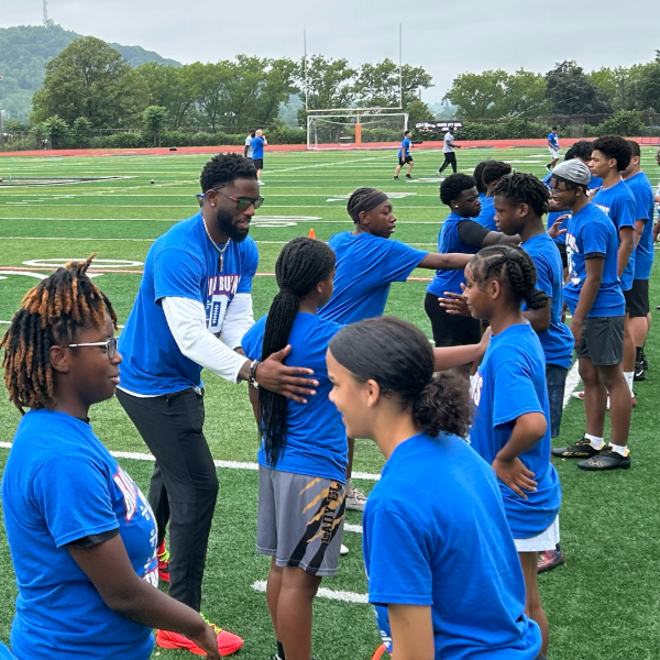 Football coach and kids in blue shirts huddle playfully on a sports field. Smiles and camaraderie convey a joyful, supportive atmosphere.