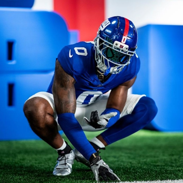 Football player Brian Burns in blue uniform crouches on turf, wearing a helmet and reflective visor. The expression is intense, with a bold backdrop.