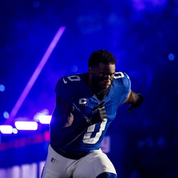 Football player Brian Burns in a blue jersey runs with determination against a dramatic, purple-lit stadium background, conveying energy and focus.