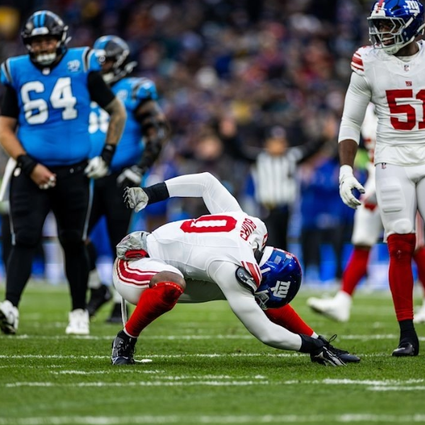 Football player Brian Burns in a white and red uniform bends over on the field, appearing tense, surrounded by others in blue and black. The scene conveys intensity and focus.
