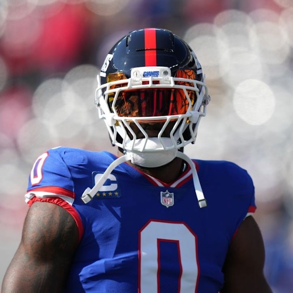 Football player Brian Burns in a blue and red jersey with number 0, wearing a shiny helmet and tinted visor, stands on the field, exuding focus and readiness.