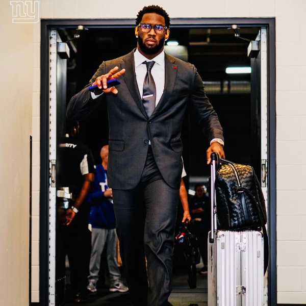 Founder Brian Burns in a sharp grey suit walks confidently down a corridor, pulling a silver suitcase. He's giving a peace sign, exuding a calm, collected demeanor.