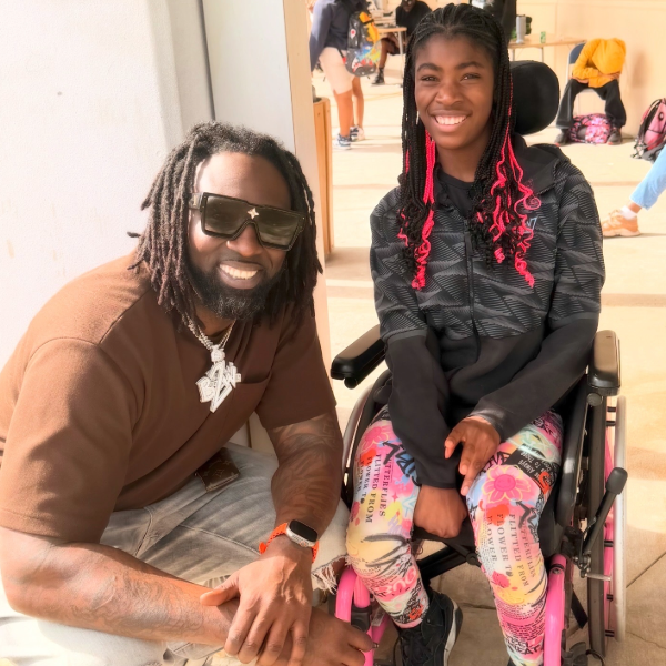 A man with braids and sunglasses kneels next to a young woman in a wheelchair, both smiling in an outdoor setting.