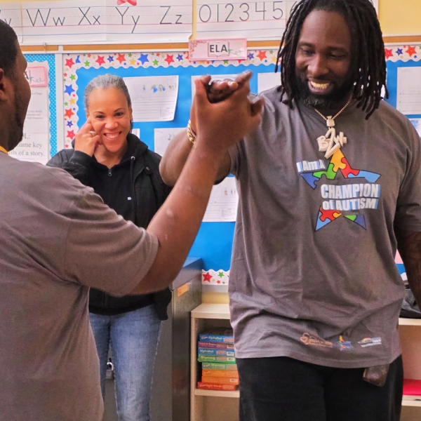Three people in a classroom, two men clasping hands and a woman smiling in the background.
