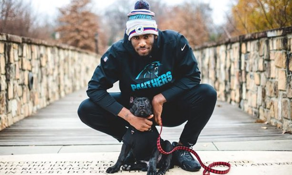 Brian Burns squatting on a walkway with a black dog, surrounded by stone walls and autumn trees.